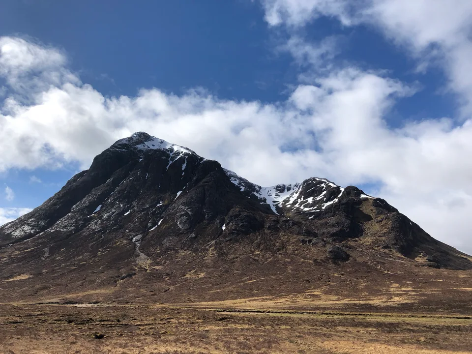 A dark rocky mountain with patches of snow under billowing white clouds.