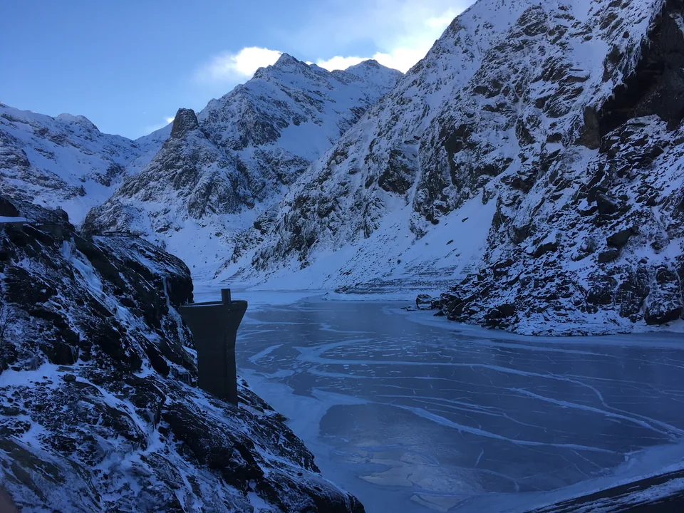 A frozen lake and a small dam below steep, snow-covered mountain walls.