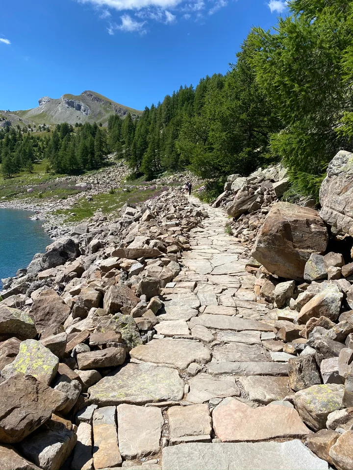 A stone path follows a blue lake between large boulders and a wooded slope.