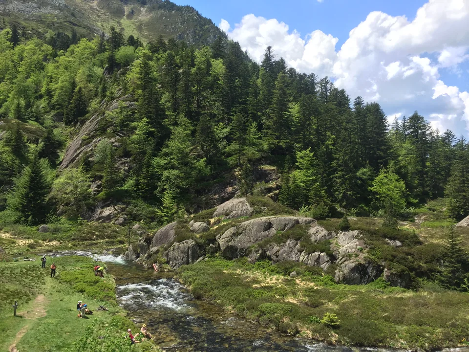Hikers beside a clear stream in a grassy clearing below a forested mountain slope.