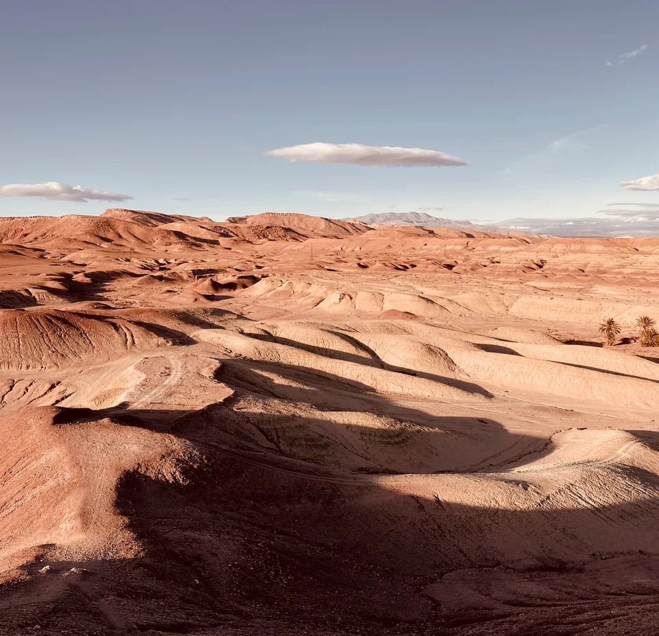 Warm reddish hills and winding folds of sand beneath a pale blue sky.