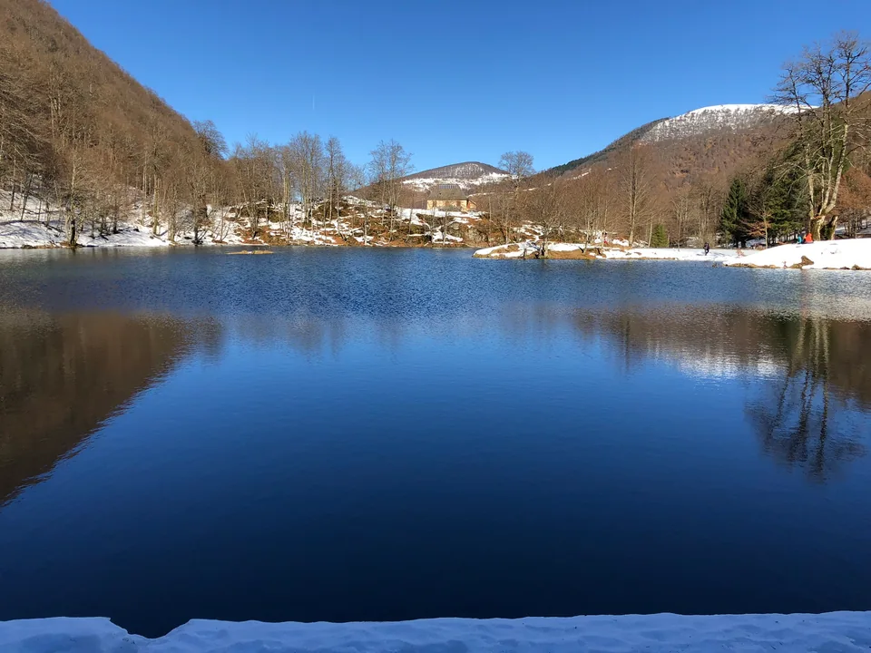 Blue lake water bordered by snowy banks and bare trees, with a building in the distance.