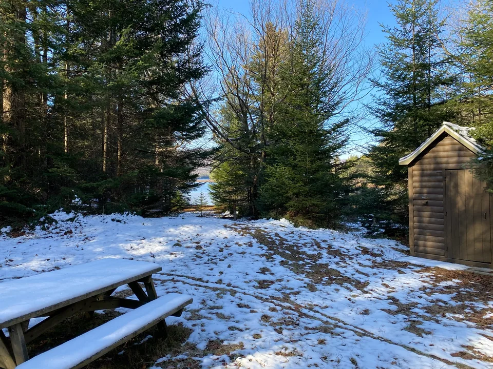 A snowy woodland clearing with a wooden hut and a picnic table.